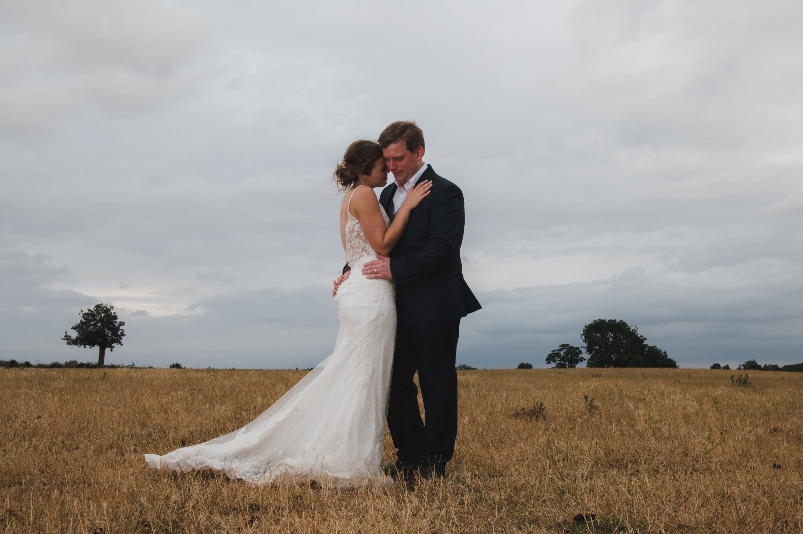 A bridal couple are embracing in the middle of a parched, grassy meadow - Bedfordshire Wedding Photographer