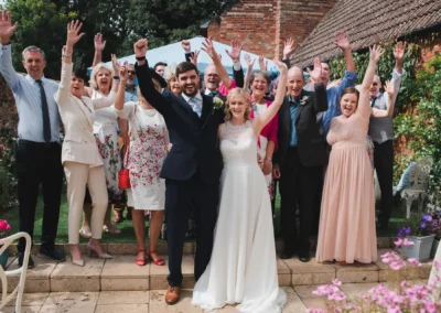 A bride and groom and their wedding party are in the grounds next to a red bricked building all with an arm raised in celebration - Event photography in Buckinghamshire