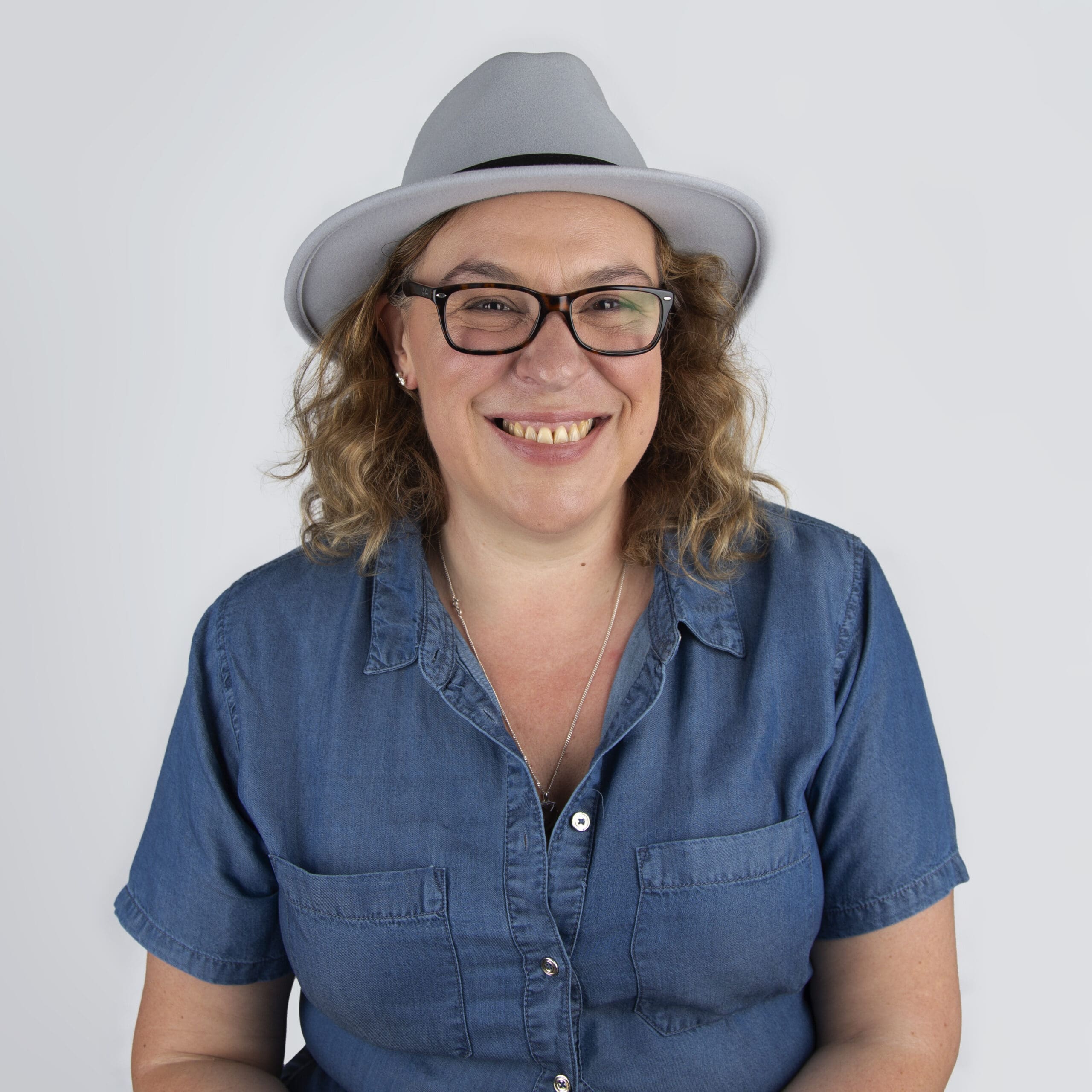 Catherine Brooks Photography - Image of Catherine a lady wearing a denim shirt with curly mouse brown hair to the shoulder and glasses. She has a grey fedora hat on her hat and black glasses and is smiling