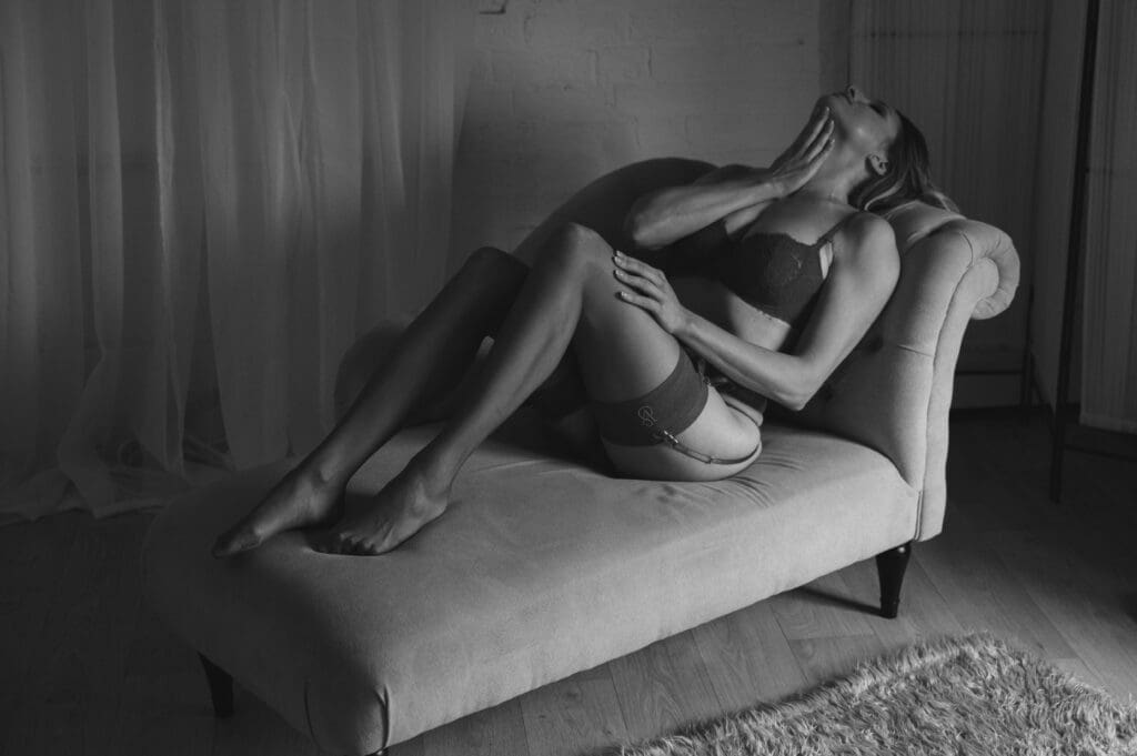 things to do before your boudoir session - A B&W image of a lady in sexy underwear and lingerie lying on a chaise long with her knees slightly up and her right hand stroking her neck