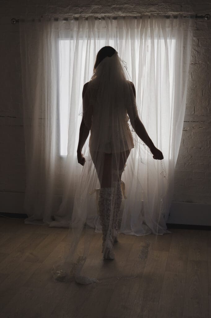 Unleash Your Inner Goddess - a lady in white bridal lingerie and a veil is stood with her back to the camera facing a net covered window in soft lighting