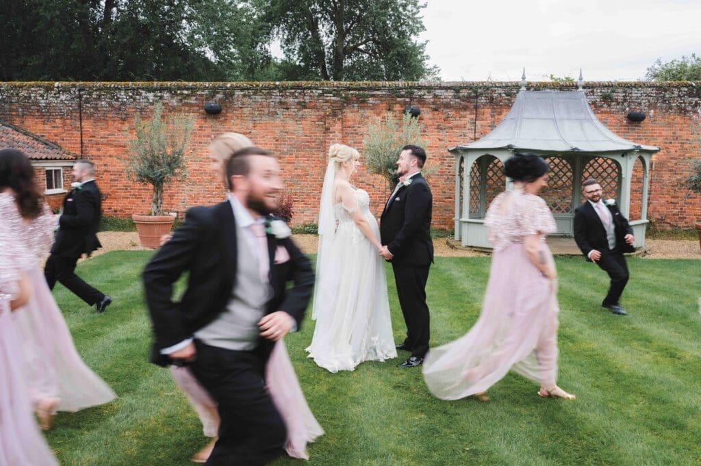 Bride & Groom standing in the pavilion garden in Braxted Park. Groomsmen and Bridesmaids are running around them